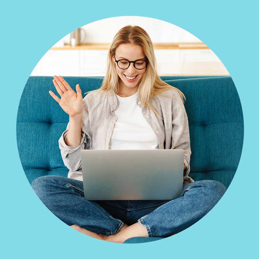 Eine Frau winkt mit der Hand und schaut währenddessen auf dem Notebook auf ihrem Schoss. Es wirkt als würde sie digital telefonieren ohne Headset.