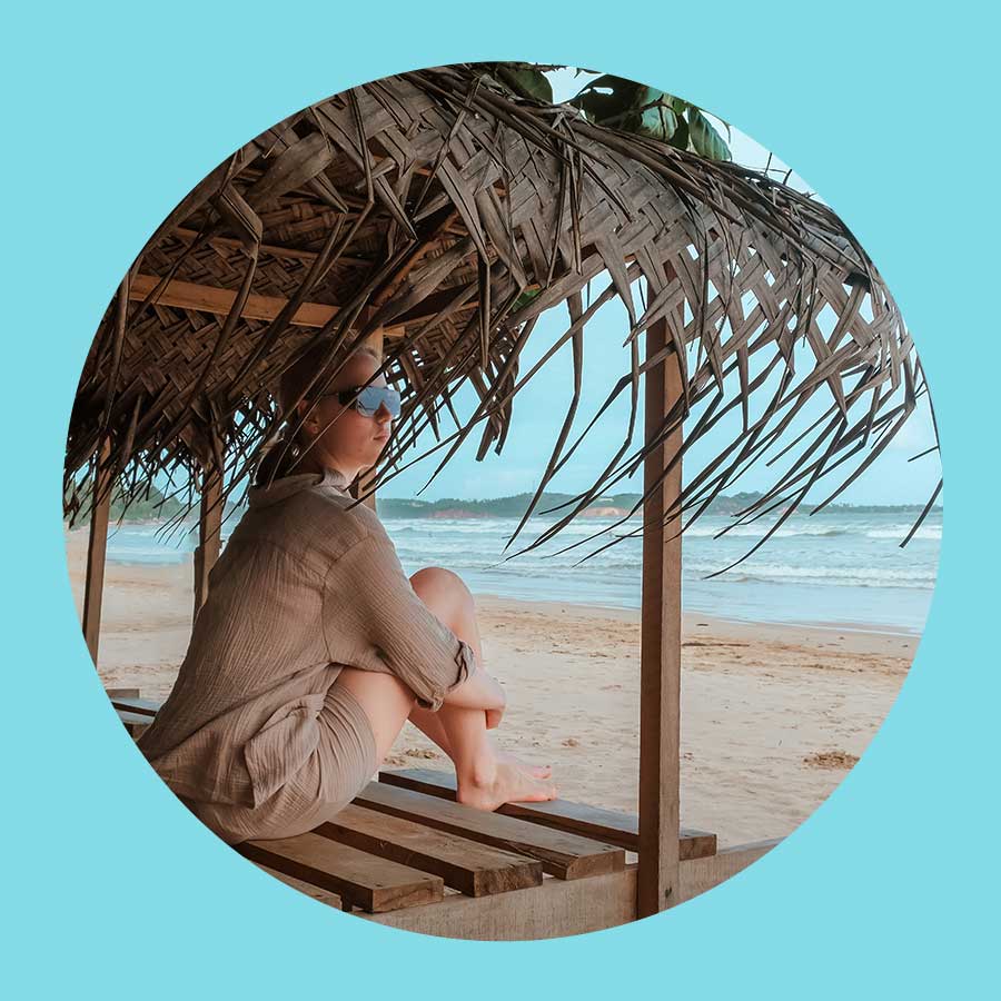 Eine Frau sitzt am Strand auf einem Sonnenschutz, der mit Palmen bedeckt ist.